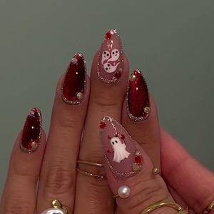May include: Almond-shaped press-on nails featuring a mix of deep red glitter polish and matte nude bases. The design includes white ghost illustrations, small red devil horns, and accents of red and clear rhinestones along the cuticle lines.