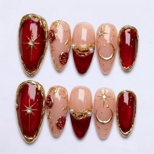 May include: A set of ten almond-shaped artificial nails. The nails feature a variety of designs, including a deep red with a gold starburst, nude with gold roses, and nude with gold crescent moons and stars. Each nail is adorned with gold accents.