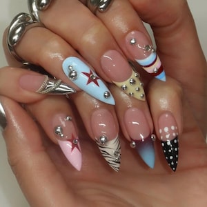 May include: A close-up of a hand with long, pointed, manicured nails. Each nail features a different design, including light pink, light blue, and black with white polka dots. Some nails have silver embellishments and star designs.