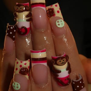 Puede incluir: Uñas postizas largas de forma cuadrada con base de manicura francesa y adornos en 3D. Los diseños incluyen figuras de monos marrones, puntas de color crema con lunares rojos, puntas marrones con puntos blancos y pequeños botones verde claro.