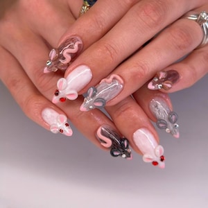May include: Nail art featuring three-dimensional sculpted mouse figures on long nails. The designs include white, grey, and brown mice with pink accents and small red or silver gemstone eyes. Some nails have a solid white base, while others are clear.