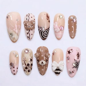 Op de afbeelding: Een set van tien amandelvormige kunstnagels in beige, bruine en roze tinten. De ontwerpen bevatten 3D-beertjes, witte strikken, bloemmotieven, muzieknoten, sterren, stippen en spiralen, geaccentueerd met kleine doorzichtige en roze strass-steentjes.
