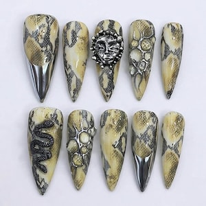 May include: A set of ten stiletto-shaped press-on nails featuring a beige and black snakeskin pattern. The nails are decorated with silver metallic tips, a three-dimensional silver sun face charm, a black snake figure, and circular silver accents.