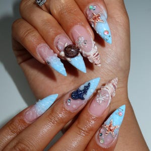 May include: Stiletto-shaped nails featuring a light blue and clear gradient base with 3D ocean-themed embellishments. Decorations include miniature sea turtles, starfish, whale sharks, and seashells, accented with small iridescent beads and clear rhinestones.