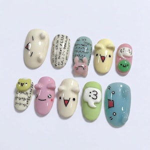 May include: A collection of eleven painted artificial nails with cartoon designs. The nails are painted in various colours, including white, pink, yellow, green, and blue. Some nails feature faces, while others have text and symbols.