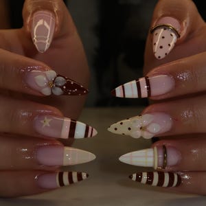Op de afbeelding: Lange amandelvormige nagels met diverse ontwerpen, waaronder bruine en witte strepen, stippen, ruitpatronen en driedimensionale witte bloemaccenten met gouden harten. Sommige nagels zijn versierd met dunne gouden metalen ringetjes.