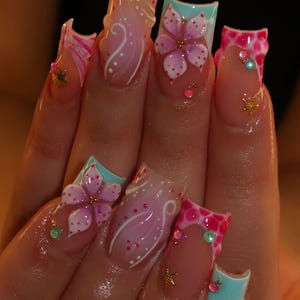 Puede incluir: Uñas postizas de forma cuadrada con una mezcla de diseños que incluyen patrones florales en rosa y blanco, líneas blancas en espiral y texturas de puntos en rosa brillante. Las uñas están decoradas con pequeños dijes de estrellas de mar doradas y pedrería.