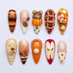 May include: A set of ten almond-shaped press-on nails featuring autumn-themed 3D designs. Patterns include brown and orange stripes, coffee beans, a bear with a pumpkin, cherry pie, apple slices, a swirl, maple leaves, pumpkin pie, and polka dots.