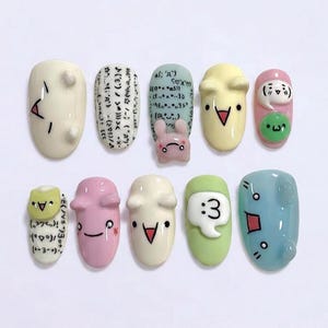 May include: Ten artificial nails with diverse designs and colours. The nails are painted in white, pink, yellow, green, and blue. Some nails feature cartoon faces, symbols, and text. Designs include a bunny, a speech bubble with the number 3, and geometric shapes.