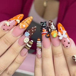 May include: Almond-shaped nails featuring a clear base with intricate 3D nail art. Designs include orange and brown polka dots, stripes, a black cat figurine, small white buttons, a white bow, and a miniature orange slice charm on the nail tips.