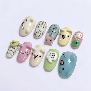 May include: A set of ten decorative nail art designs. Each nail showcases a unique design, including cartoon faces, mathematical symbols, and a small bunny. The nails are painted in pastel shades of pink, green, and blue.