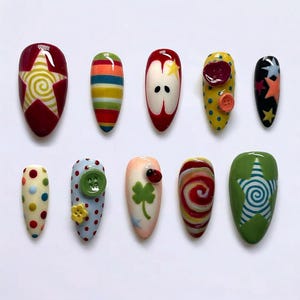 Könnte beinhalten: Eine Sammlung von zehn künstlichen Fingernägeln mit einzigartigen Designs. Jeder Nagel hat ein anderes Design, darunter Sterne, Streifen, Äpfel, Punkte und ein vierblättriges Kleeblatt. Die Nägel sind in verschiedenen Farben wie Rot, Gelb, Grün und Blau bemalt.