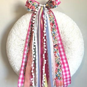 May include: A white wreath with a textured surface, adorned with a large bow and colorful ribbons. The bow features a floral pattern and pink gingham, with ribbons in various colors and textures, including lace and pom-poms.