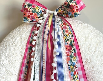 May include: A decorative bow featuring a pink gingham pattern, floral print, and various ribbons. White pom-poms and lace trim add detail. The bow is attached to a fluffy white surface.