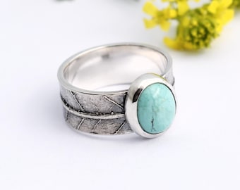 Puede incluir: Un anillo de plata con diseño de hoja y una piedra ovalada turquesa. El anillo se encuentra sobre un fondo blanco con flores amarillas y hojas verdes en el fondo. Una joya.