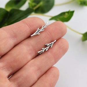 Puede incluir: Un par de pendientes de plata con forma de ramas delicadas con peque&ntilde;as hojas. Los pendientes se muestran en una mano, con un pendiente en cada dedo.