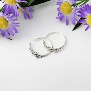 May include: A pair of silver hoop earrings with a delicate twig design. The earrings are on a white background.