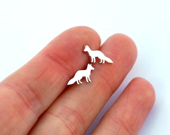 Peut inclure: Deux petites boucles d'oreilles argentées en forme de renard. Les boucles d'oreilles ont un design simple, avec une silhouette de renard debout, la queue tendue. Les boucles d'oreilles sont présentées sur le doigt d'une personne.