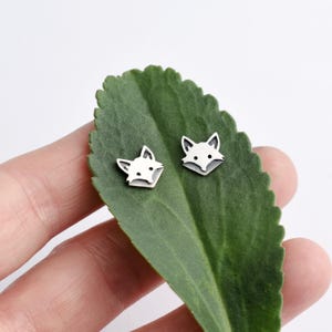 May include: A pair of silver fox head stud earrings on a green leaf. The fox heads are facing forward with their ears pointed up.