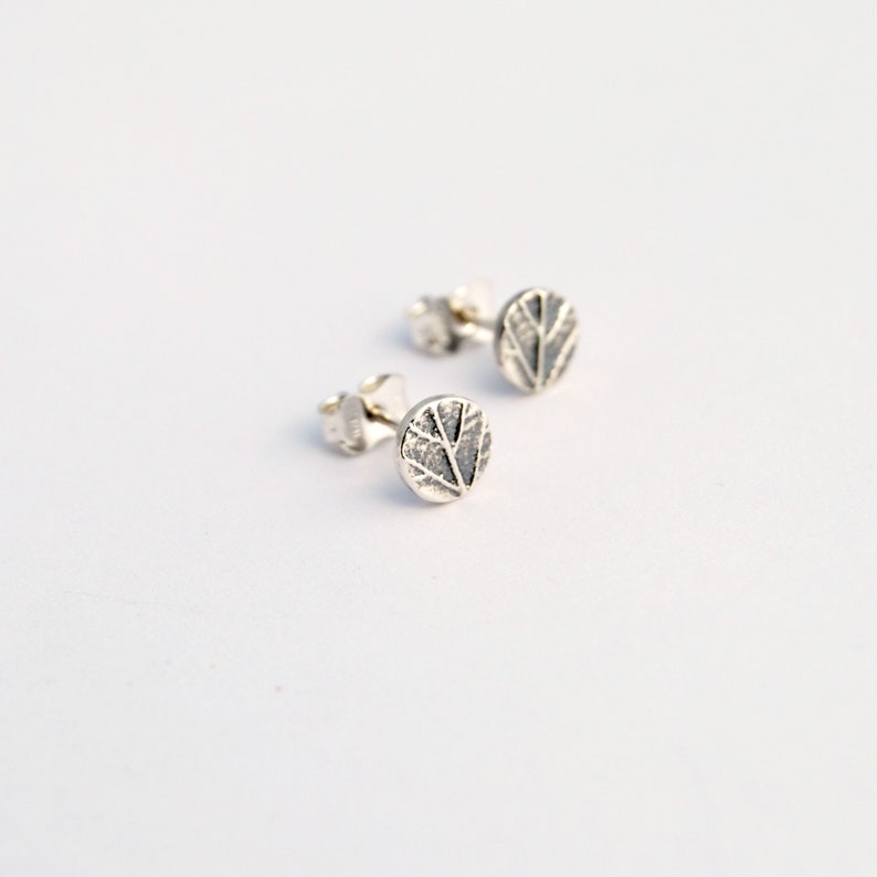Puede incluir: Un par de pendientes de bot&oacute;n de plata con un dise&ntilde;o de hoja texturizada.