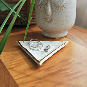 Puede incluir: Un plato triangular de cer&aacute;mica blanca con un borde dorado, utilizado para guardar joyas. El plato est&aacute; sobre una superficie de madera y contiene dos pendientes de aro de plata y un par de pendientes de bot&oacute;n de plata.