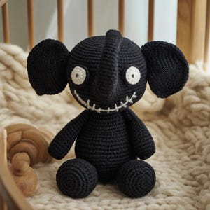 May include: A black crocheted elephant plush toy with large ears, a trunk, and white stitched eyes and mouth. The elephant is sitting on a cream-colored knitted blanket. The toy has a whimsical, slightly spooky design.
