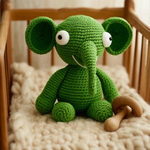 May include: A handmade green crochet elephant with large ears and white eyes. The elephant is sitting on a cream-colored blanket, with a wooden mushroom toy nearby. The toy is designed for babies and toddlers.