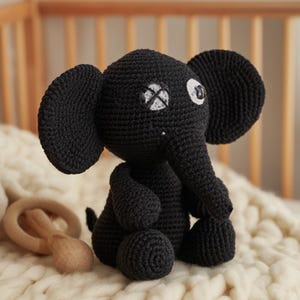 May include: A black crocheted elephant toy with large ears, a trunk, and a small tail. The elephant has white accents around one eye and a cross-shaped design on the other. A wooden teething ring is in the background.