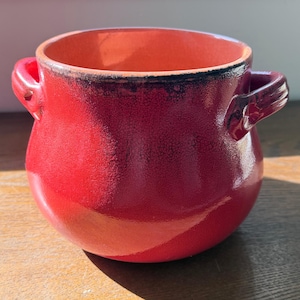May include: A round ceramic pot with two side handles, featuring a vibrant red glaze with a subtle crackle texture and dark, charred-looking edges around the rim and handles. The interior of the pot is a smooth, lighter shade of orange-red.