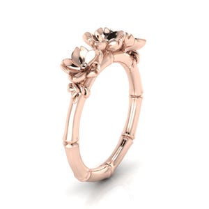 May include: A rose gold ring featuring a band designed to resemble bamboo segments. The top of the ring is adorned with three sculpted flower blossoms, each with delicate petals, positioned along the upper curve of the bamboo-textured metal band.
