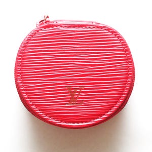 May include: A round, bright red coin purse featuring a textured, wavy surface pattern. The front displays a debossed LV logo in the center. The item is secured with a matching red zipper that runs along the perimeter of the circular structure.