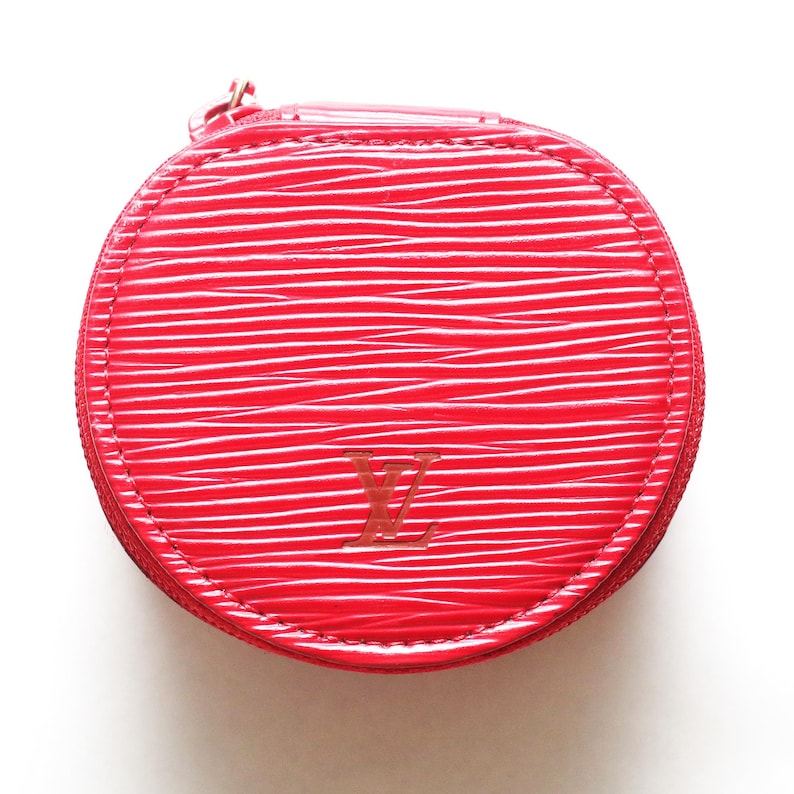 May include: A round, bright red coin purse featuring a textured, wavy surface pattern. The front displays a debossed LV logo in the center. The item is secured with a matching red zipper that runs along the perimeter of the circular structure.