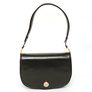 May include: A black leather shoulder bag with a smooth finish, featuring a curved flap closure and a gold-toned circular metal emblem on the front. The bag includes a single adjustable shoulder strap attached with gold-toned hardware.