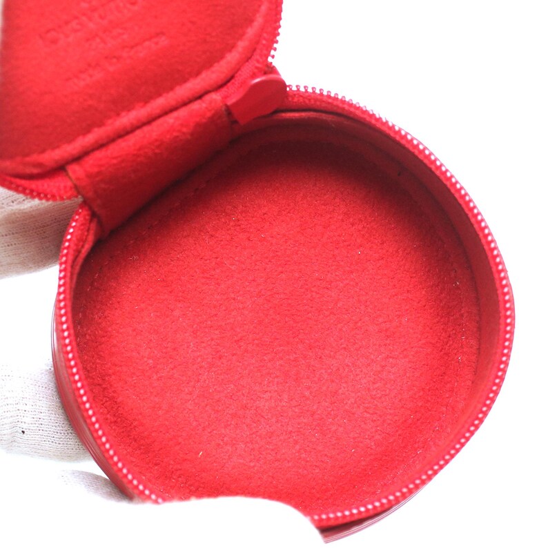 May include: A round, bright red zippered case with a matching red interior lining. The inside of the lid features embossed text that reads Louis Vuitton Paris, made in France. The case is circular in shape and features a zipper closure.