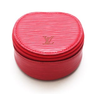 May include: A round, bright red jewelry case featuring a textured, wavy surface pattern. The top lid displays a centered, embossed LV logo. The case is secured with a matching red zipper that runs along the perimeter, separating the lid from the base.