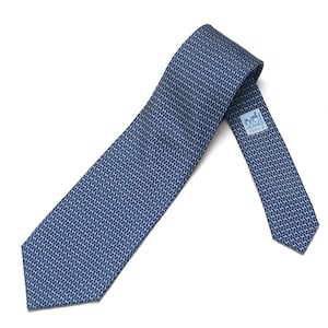May include: A blue necktie featuring a repeating white geometric pattern of small H shapes. The tie includes a light blue label on the back with the text HERMES PARIS and a graphic of a horse and carriage, indicating the brand and origin.