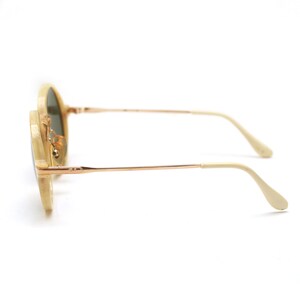 May include: Round sunglasses with a light beige frame and gold-colored metal arms. The lenses are a dark green color. The arms curve down at the ends. The sunglasses are set against a white background.