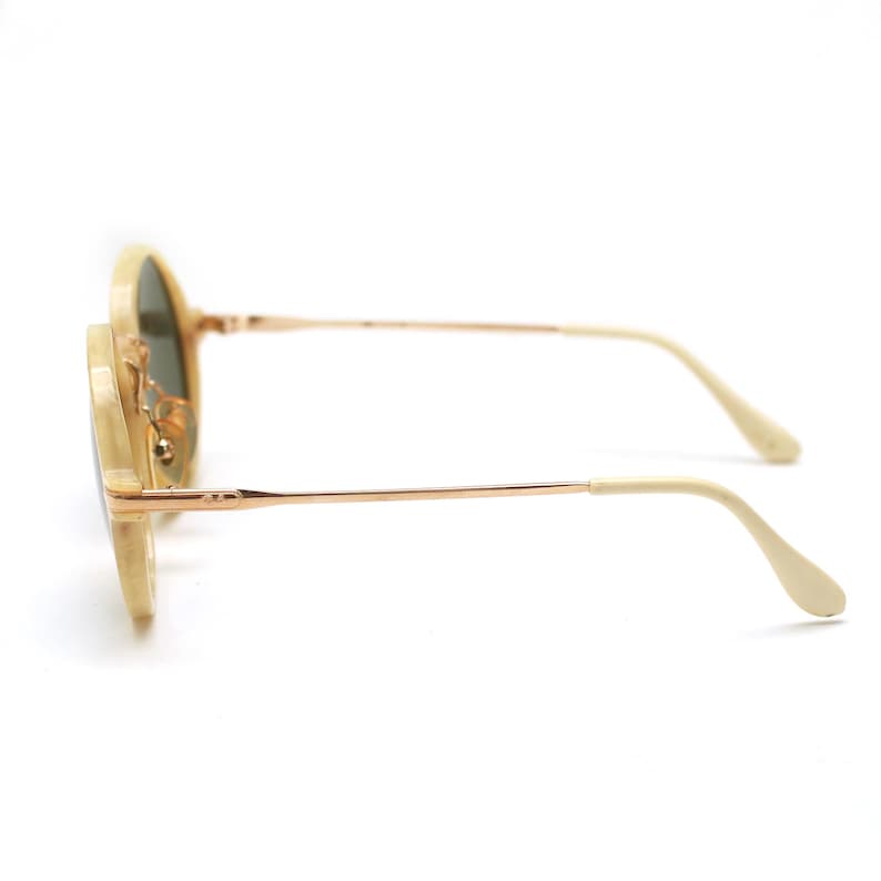May include: Round sunglasses with a light beige frame and gold-colored metal arms. The lenses are a dark green color. The arms curve down at the ends. The sunglasses are set against a white background.