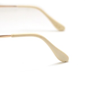 May include: Close-up of eyeglasses with a gold-colored frame and off-white temple tips. The glasses are set against a white background, highlighting the details of the frame and arms.