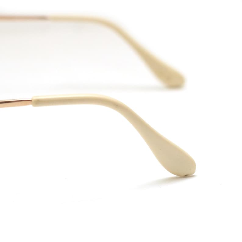 May include: Close-up of eyeglasses with a gold-colored frame and off-white temple tips. The glasses are set against a white background, highlighting the details of the frame and arms.