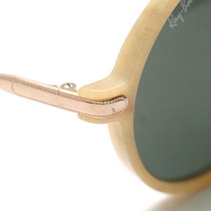 May include: Close-up of a pair of sunglasses with a light-colored frame and green lenses. The arm of the glasses is a rose gold color, with a hinge connecting to the frame. The Ray-Ban logo is visible on the lens.