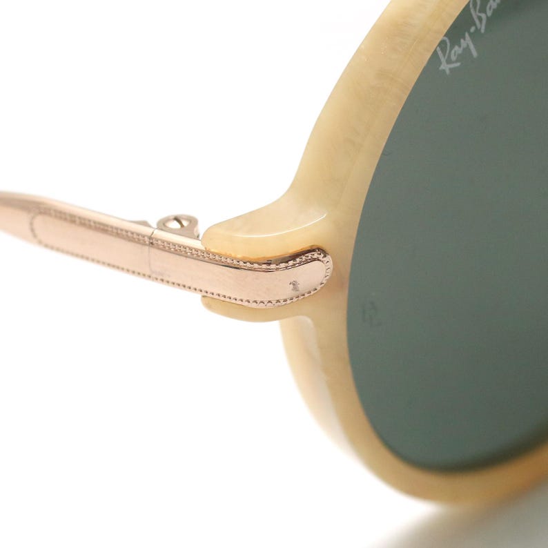 May include: Close-up of a pair of sunglasses with a light-colored frame and green lenses. The arm of the glasses is a rose gold color, with a hinge connecting to the frame. The Ray-Ban logo is visible on the lens.