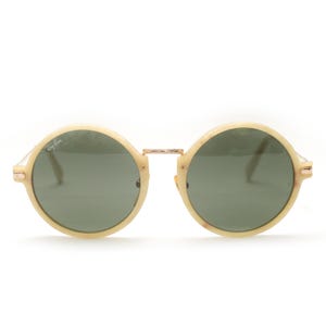 May include: Round sunglasses with a cream-colored frame and dark green lenses. The sunglasses have a gold-colored bridge and hinges. The Ray-Ban logo is visible on the left lens. A stylish accessory for eye protection.