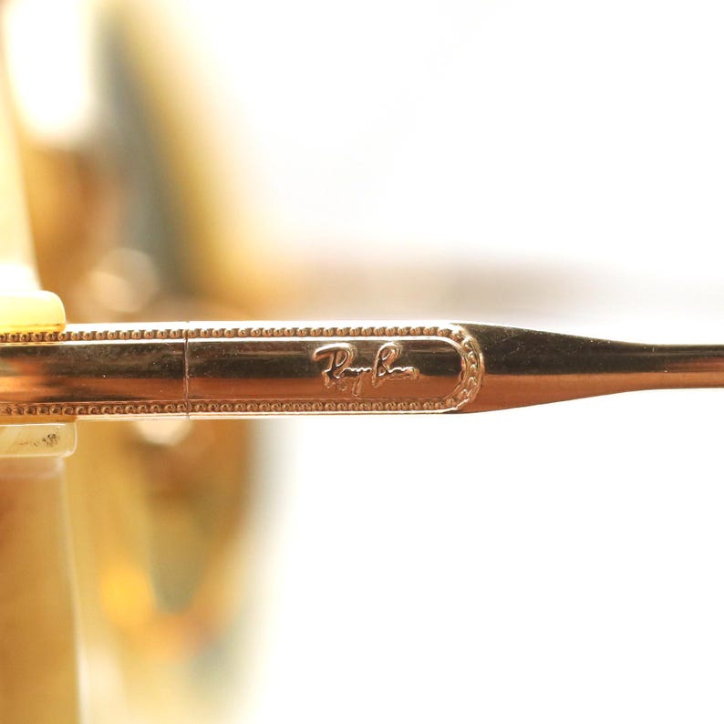 May include: Close-up of a gold-colored Ray-Ban eyeglass arm. The arm features the Ray-Ban logo in a rectangular, ornate frame. The arm is thin and tapers towards the end, with a detailed, textured design along the edges.