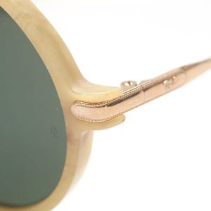 May include: Close-up of sunglasses with a light beige frame and dark green lenses. The temples are a gold color with a decorative design. The sunglasses are set against a white background.