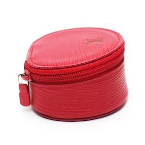May include: A round, bright red zippered pouch featuring a textured, wavy line pattern across the exterior. The top lid displays a small embossed logo, and a matching red leather zipper pull with an embossed logo is attached to the metal zipper.