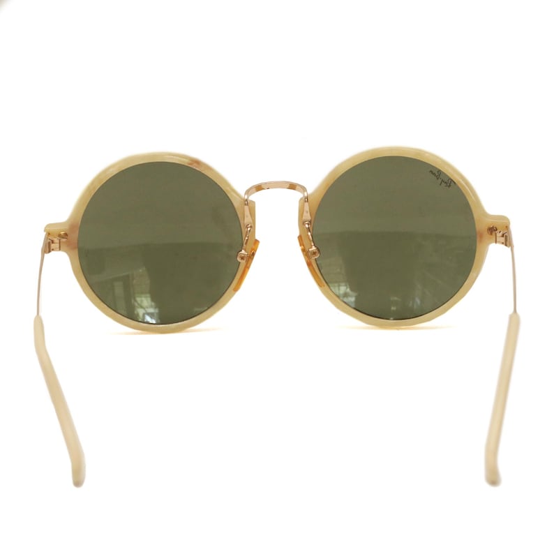 May include: Round sunglasses with a light beige frame and green lenses. The glasses have a gold-colored bridge and temples, and the arms are a light beige color. The lenses have a slight reflection.