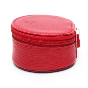 May include: A round, bright red coin purse featuring a textured, wavy line pattern on the exterior. The accessory has a matching red zipper closure that runs along the top edge and a small, embossed logo on the upper surface of the lid.