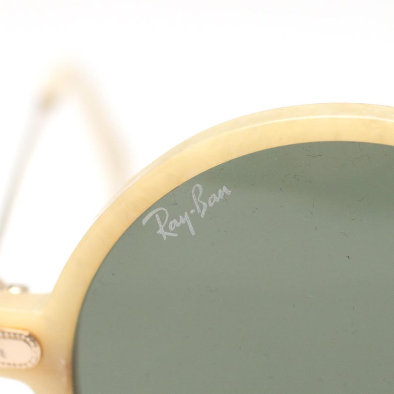 May include: Close-up of Ray-Ban sunglasses. The sunglasses have a light cream-colored frame and green lenses. The Ray-Ban logo is visible on the lens. The sunglasses are a classic style.