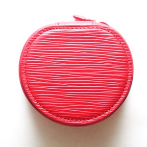 May include: A round, bright red jewelry case featuring a textured, horizontal wavy line pattern across its surface. The case is constructed with a stitched border around the circular lid and includes a small zipper pull at the top edge.